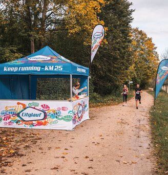 The blue and white event tent serves as a supply station for a marathon. 2 runners are passing the event tent.